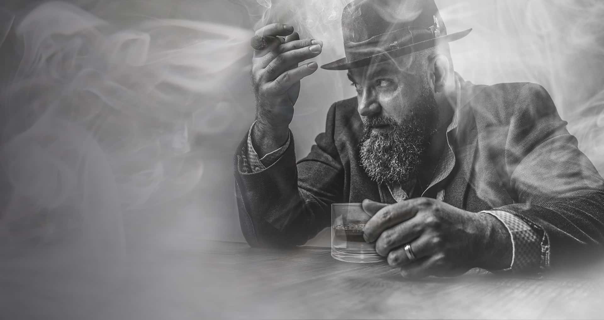 Man sitting at the bar in a suit and fedora having a drink and smoking a cigar.