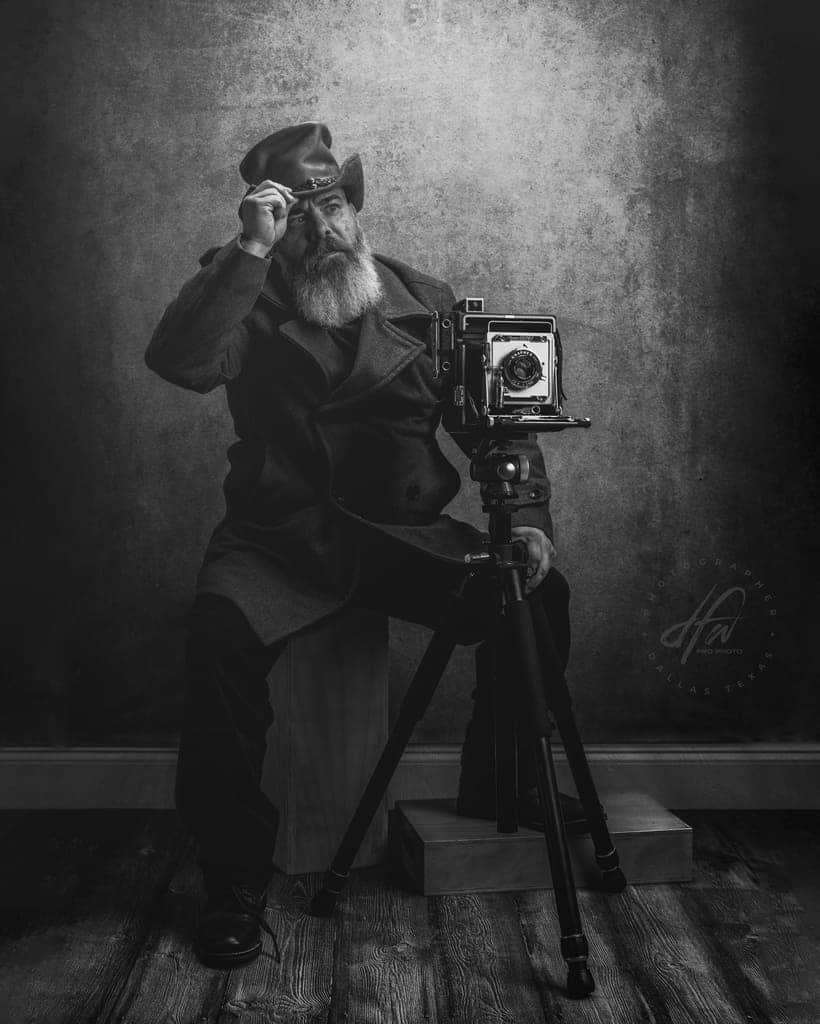 Man sitting on apple box behind and old camera wearing a top hat and peacoat, tipping his hat.