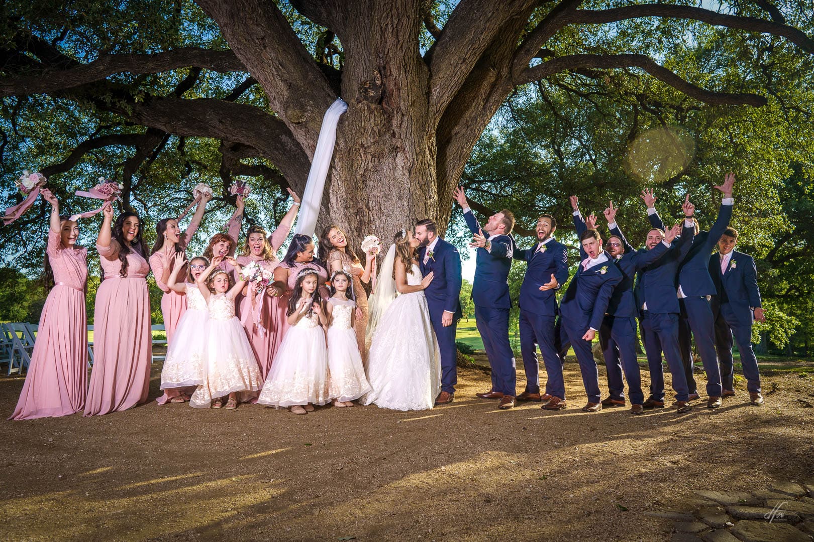 Newlyweds kiss surrounded by wedding party under a huge tree.