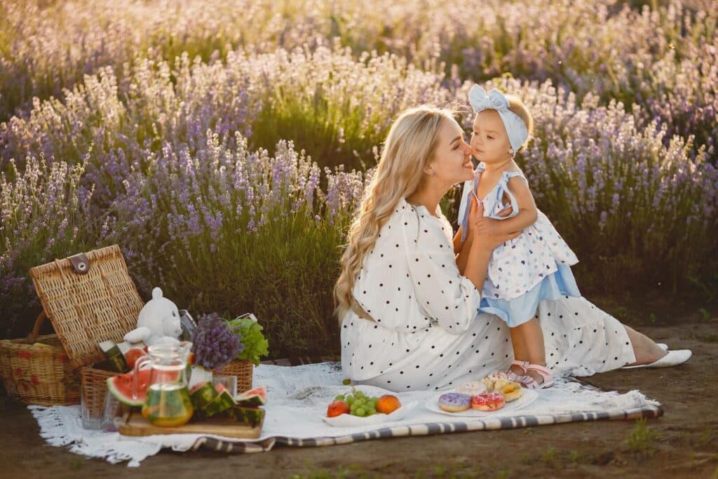 Mom hold toddler on a picnic blanket with flowers in the background.
