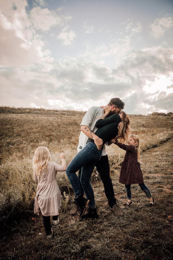 Couple outside kissing with their two young girls playing around them.