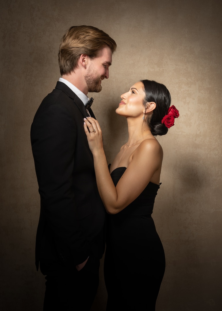 Couple in black formal wear posing for photographer in front of a canvas backdrop.